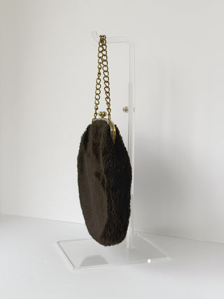 Side profile view of vintage brown faux fur clutch showing textured plush surface and gold chain strap hanging from clear display stand