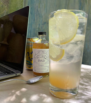 MOTHER shrub Ginger bottle next to tall glass of lemonade with lemon slice garnish on desk beside laptop computer