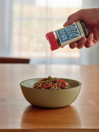 Hand holding Woon Sea Moss Seasoning jar tilted over bowl of soba noodles with pickled vegetables demonstrating product use