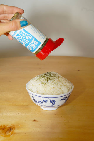 Hand sprinkling Woon Sea Moss Seasoning from tilted jar with red cap onto white rice in blue and white ceramic bowl