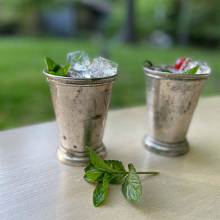 Two silver mint julep cups with ice and mint garnish, featuring MOTHER Black Cherry shrub on outdoor table