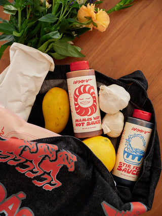 Woon hot sauce bottles in black checkered tote bag with mango, garlic, lemons and flowers