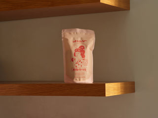 Woon Hibiscus tea package resting on wooden shelf against neutral wall showing minimalist product styling and pink packaging