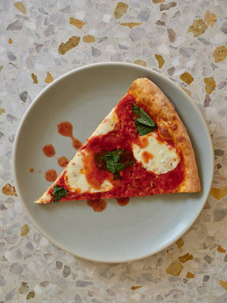 Slice of margherita pizza with basil on white plate drizzled with Woon hot sauce on terrazzo countertop