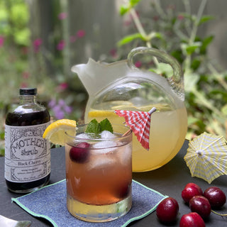 MOTHER Black Cherry shrub bottle with fresh cherries, cocktail with checkered straw, and pitcher of lemonade on outdoor garden table
