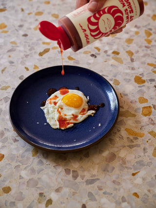Hand squeezing Woon Mama's Way hot sauce bottle over fried egg on blue plate showing sauce dripping