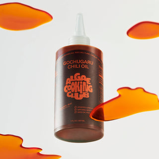 Bottle of Gochugaru Chili Oil by Algae Cooking Club with white squeeze cap against light background with orange chili oil splashes