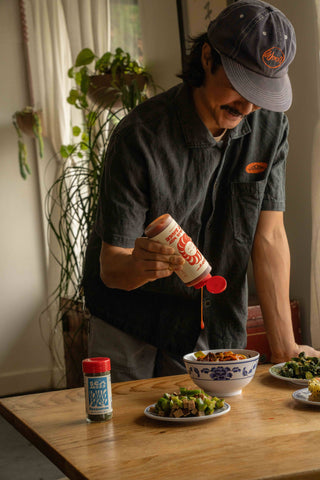 Person in Woon branded cap and dark shirt pouring Mama's Way hot sauce over bowl of food on wooden table