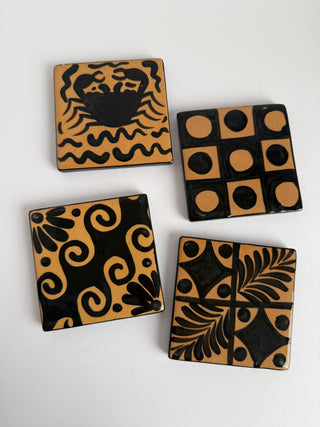 Four vintage Honduran folk art coasters laid flat showing hand-painted black and tan designs including crab, checkerboard, spiral, and fern leaf patterns