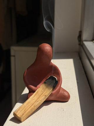 Vintage terracotta ceramic figure with rounded head and body holding a wooden palo santo stick with visible smoke rising, photographed on white surface in natural sunlight