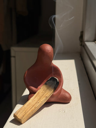 Vintage terracotta ceramic figure with organic sculptural form and integrated holder displaying a burnt palo santo stick with smoke trail, shown on white surface with natural lighting