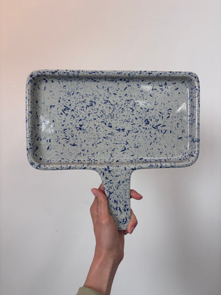Vintage blue and white speckled enamelware tray with handle held against neutral background, showing interior and top-down view