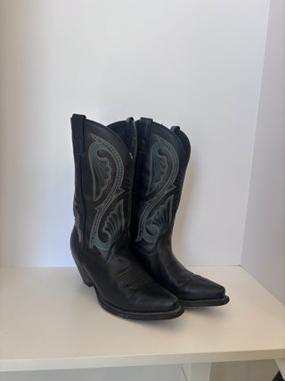 Side angle view of vintage black leather cowboy boots with ornate blue stitching on white shelf