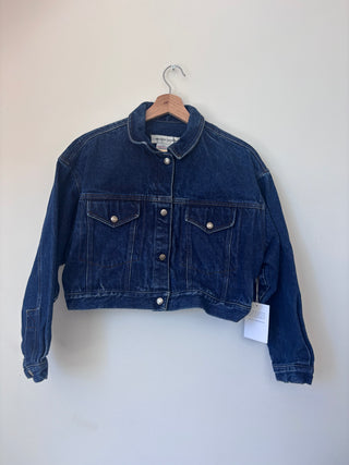 Front view of vintage Calvin Klein Sport cropped denim jacket on wooden hanger against neutral wall, showing snap button closure and chest pockets
