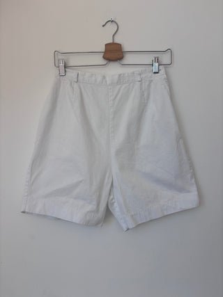 Vintage white cotton tailored shorts hanging on wooden hanger against neutral wall, front view showing pleated detailing and cuffed hem