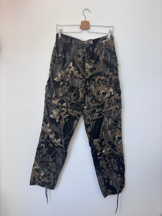 Vintage tree bark camo cargo pants hanging on wooden hanger against white wall, front view