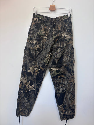 Vintage tree bark camo cargo pants hanging on wooden hanger against white wall, back view showing multiple cargo pockets