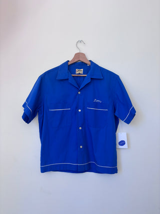 Front view of vintage royal blue Hilton work shirt with white piping and 'Don' embroidered on chest pocket, hanging on wooden hanger