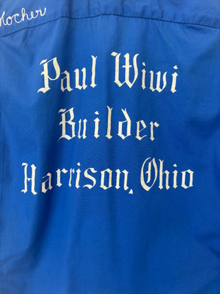 Detail view of white chain-stitch embroidery reading 'Paul Wiwi Builder Harrison, Ohio' in gothic font on blue shirt back