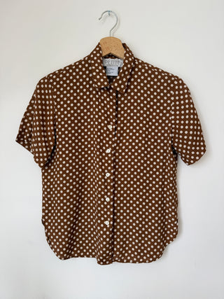 Front view of vintage brown polka dot short sleeve button-up shirt hanging on wooden hanger against white wall, showing Petite Sophisticate label