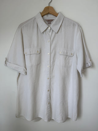 Front view of vintage cream linen button-up shirt hanging on wooden hanger against white wall, showing chest pockets and roll-tab sleeves