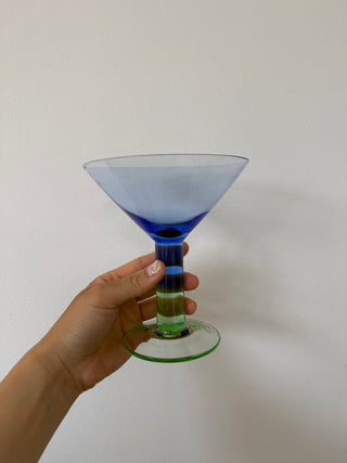 Vintage blue art glass martini coupe held by hand against white background, showing ridged multicolor stem with cobalt blue and lime green bands and circular green base