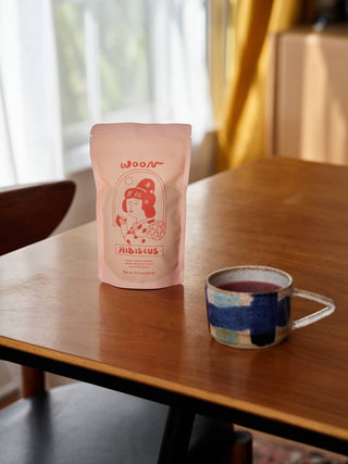 Woon Hibiscus tea pouch displayed on wooden table next to handmade ceramic mug filled with brewed red hibiscus tea in modern interior setting