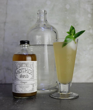 MOTHER shrub Ginger 8oz bottle with white illustrated label next to tall glass cocktail with mint garnish and clear bottle on marble surface