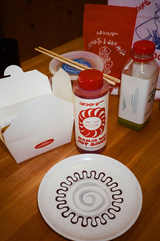 Woon Mama's Way hot sauce bottle on table with takeout boxes, chopsticks, and decorative plate with sauce design