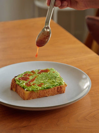 Spoon holding Woon hot sauce dripping over avocado toast on white plate on wooden cutting board