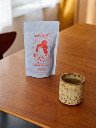 Woon Genmaicha tea pouch displayed on wooden table next to handmade ceramic cup in neutral interior setting
