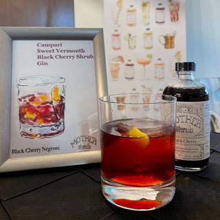 Black Cherry shrub bottle next to framed cocktail recipe card and finished drink garnished with lemon twist on dark countertop