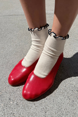 Close-up of cream ribbed sock with navy wavy ruffle trim paired with red vintage leather mary jane shoe
